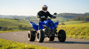 Pilote en casque sur quad sportif bleu homologué neuf, sur route goudronnée sinueuse traversant des collines verdoyantes sous un ciel clair.
