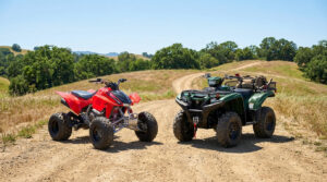 Un quad rouge sportif et un quad vert utilitaire côte à côte sur un chemin de terre en milieu rural, sous un ciel bleu.