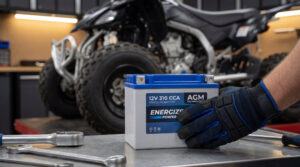 Main gantée tenant une batterie quad Energizer AGM sur un établi métallique avec des outils, et un quad en arrière-plan.