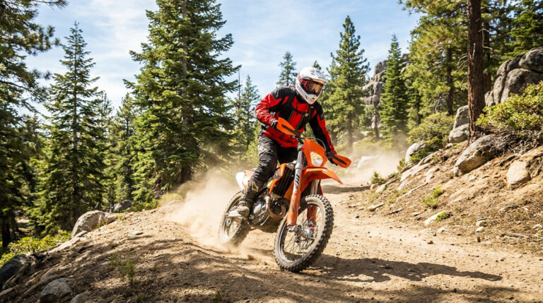 Pilote de moto enduro orange avec casque et équipement, dérapant sur un sentier forestier rocailleux et poussiéreux sous un ciel bleu.
