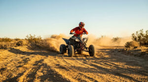 Pilote sur un quad Yamaha Raptor noir, portant un casque et une tenue rouge, traversant un paysage désertique en soulevant de la poussière.