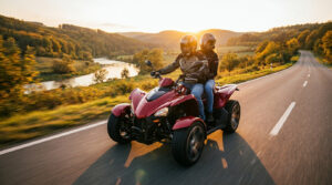 Couple casqué sur un quad rouge 2 places roulant sur une route panoramique au soleil couchant, avec rivière et collines arborées.