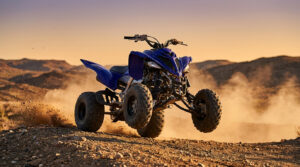 Quad Yamaha Raptor bleu en plein saut sur un terrain désertique, nuage de poussière sous les roues. Coucher de soleil en arrière-plan.