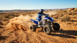 Quad Yamaha Raptor 750 bleu et blanc avec pilote masqué, soulevant un nuage de poussière sur un sentier désertique en plein soleil.
